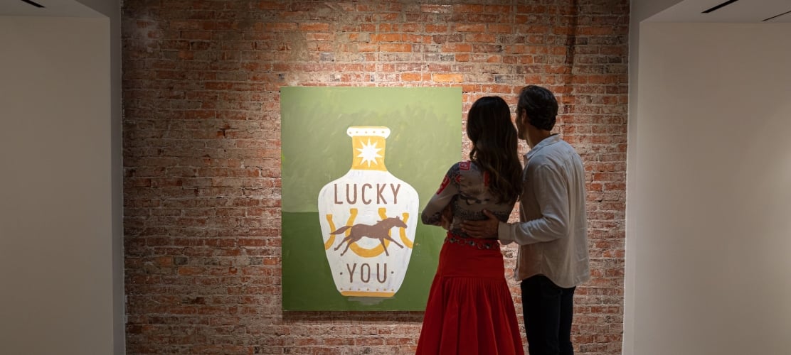 A couple observes a painting featuring a white vase with "LUCKY YOU" and a horse, displayed on a textured brick wall within an art gallery setting.