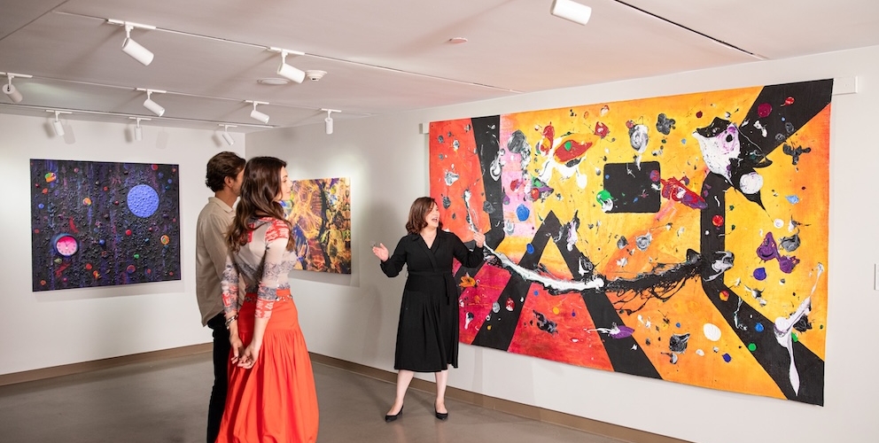 A gallerist gestures, explaining a large abstract artwork to a couple admiring it and other vibrant paintings in a modern art gallery with track lighting.