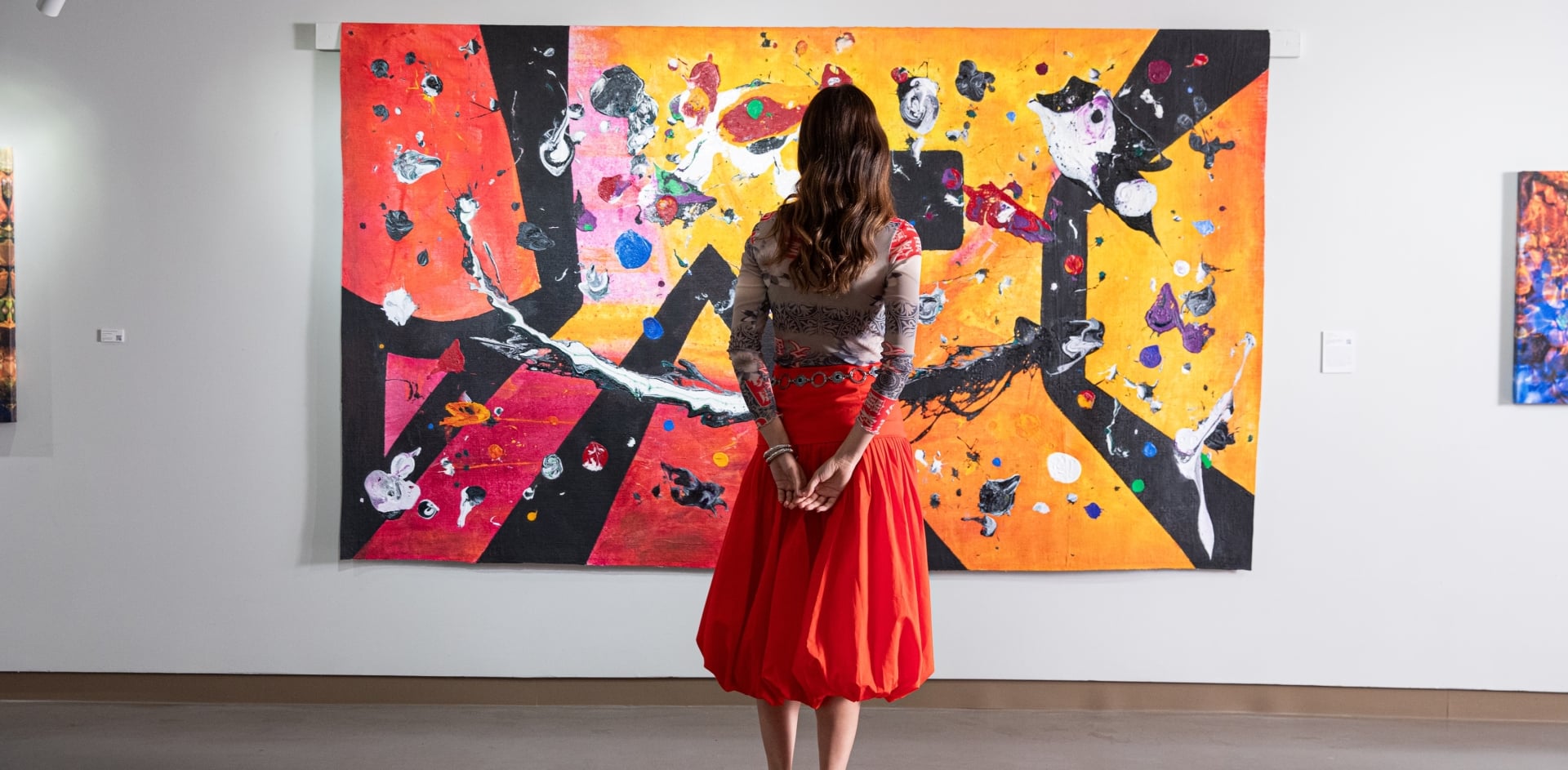 A woman with long hair wearing an orange skirt stands with her back to the camera, looking at a large, vibrant abstract painting in an art gallery.
