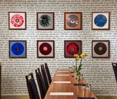 A long wooden conference table set with stationery under an art wall featuring eight framed, colorful vinyl records against text-patterned wallpaper.