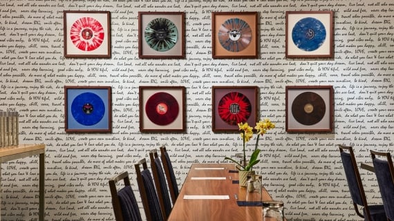 A long wooden conference table set with stationery under an art wall featuring eight framed, colorful vinyl records against text-patterned wallpaper.
