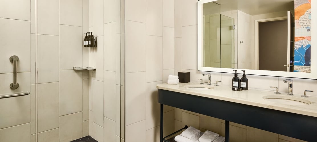 Modern hotel bathroom featuring a walk-in shower with vertical white subway tiles, a black-framed double vanity with a marble top, and a backlit mirror showing a glimpse of abstract artwork.