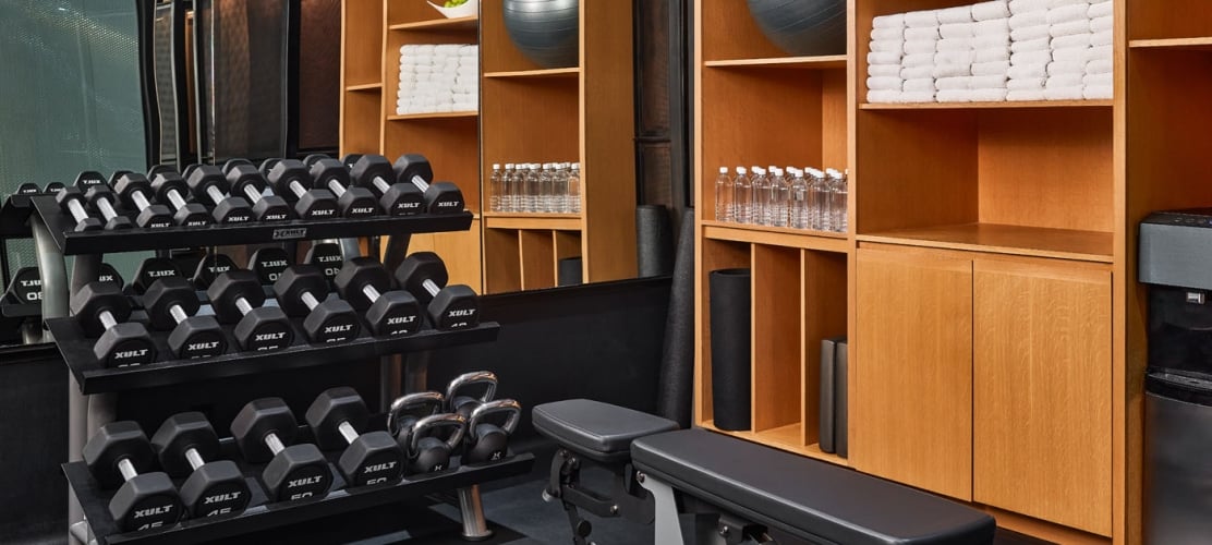 Modern hotel fitness center featuring a three-tier dumbbell rack, a workout bench, and wooden shelving stocked with rolled white towels, water bottles, and exercise balls.