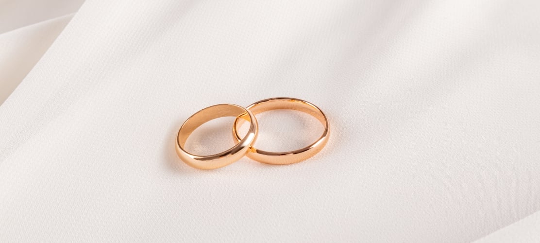 A close-up shot of two classic gold wedding bands resting together on a soft, draped white fabric background.