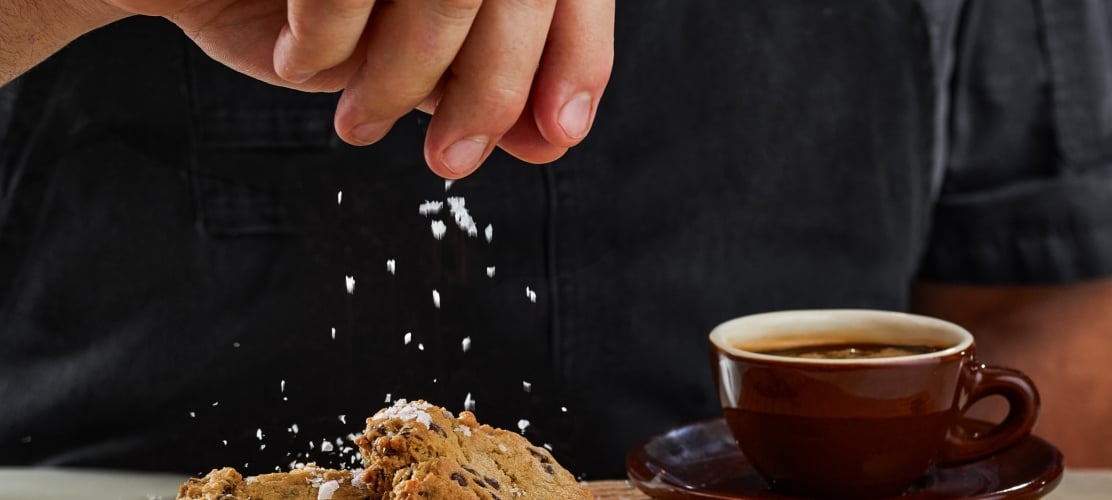 A close-up of a baker sprinkling sea salt over fresh chocolate chip cookies on a wooden board next to a cup of black coffee.