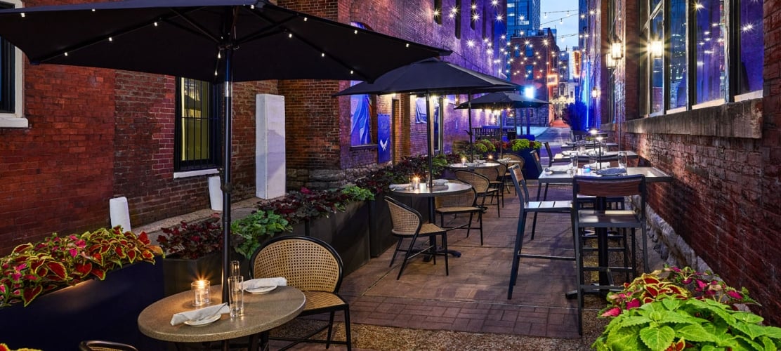 An atmospheric evening view of an outdoor restaurant patio in a historic brick alleyway, featuring tables under large black umbrellas and warm string lights.