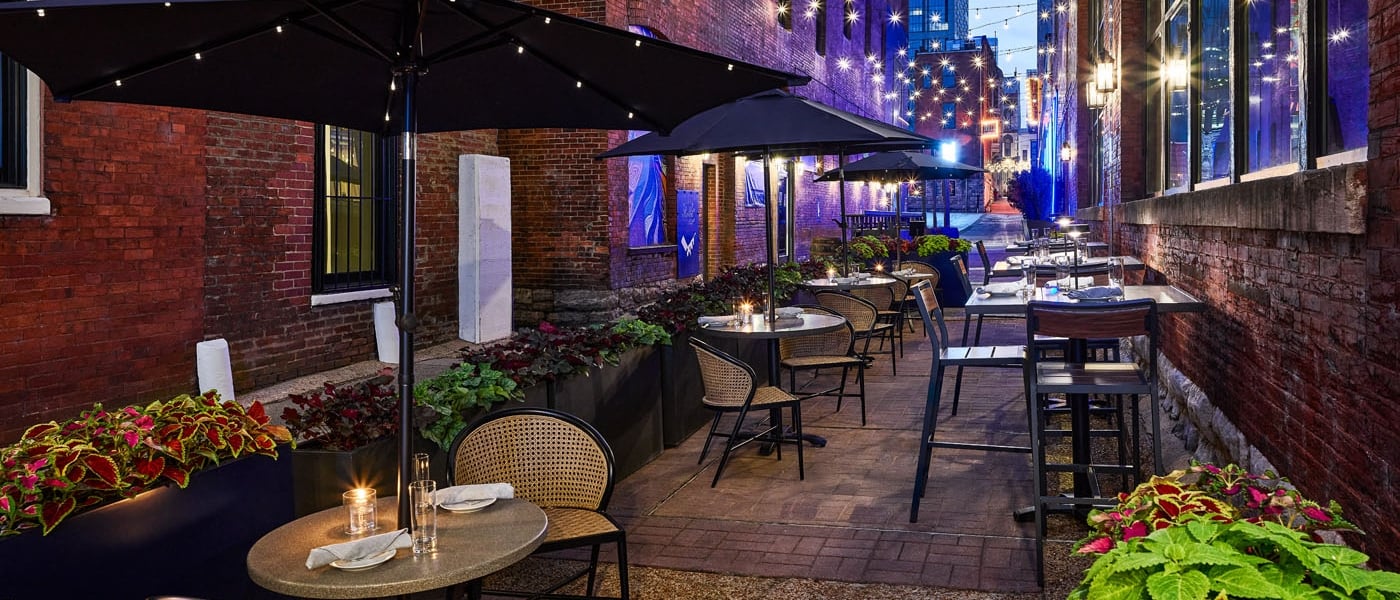 An atmospheric evening view of an outdoor restaurant patio in a historic brick alleyway, featuring tables under large black umbrellas and warm string lights.