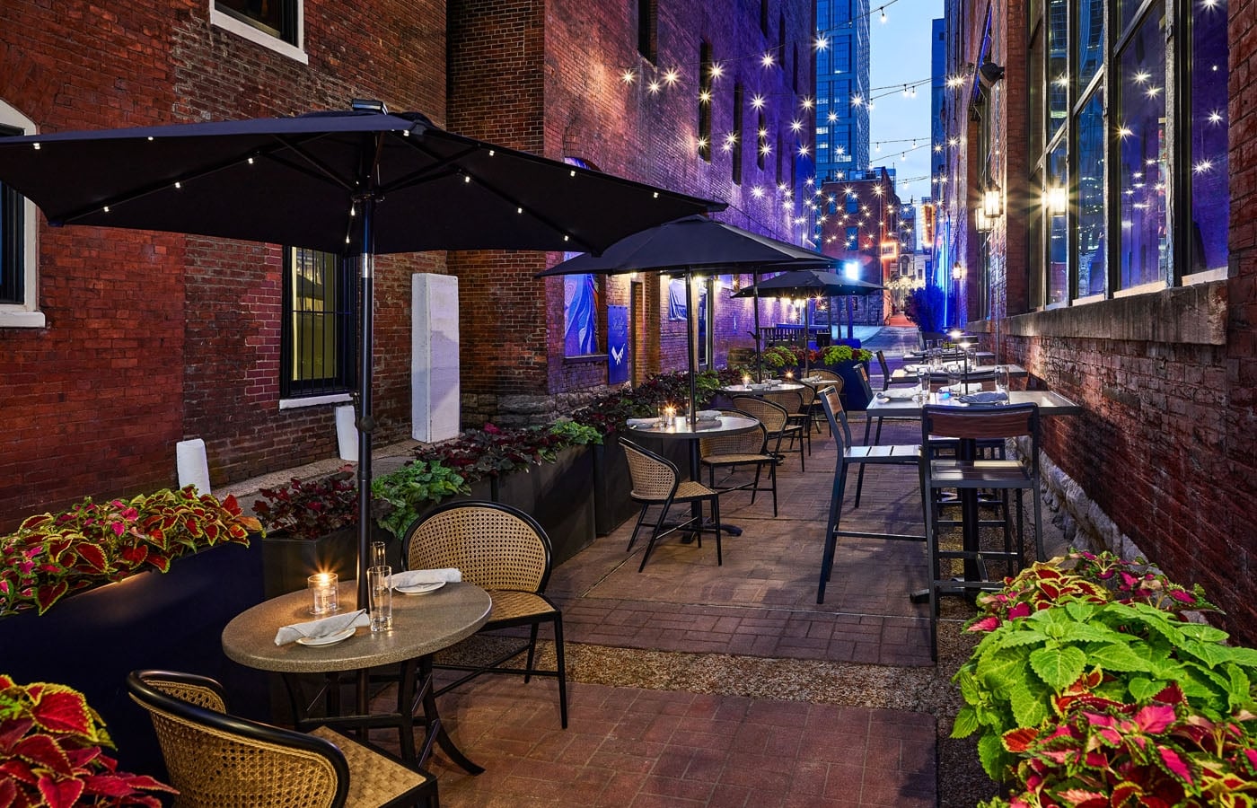 An atmospheric evening view of an outdoor restaurant patio in a historic brick alleyway, featuring tables under large black umbrellas and warm string lights.