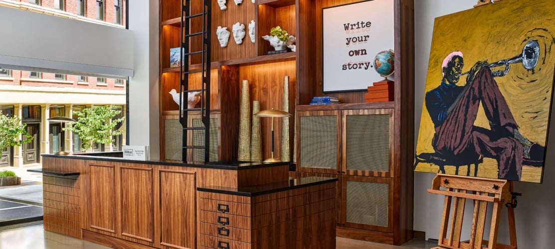 A modern luxury hotel lobby reception desk made of dark wood with a massive floor-to-ceiling wooden built-in bookshelf, a rolling library ladder, and a large oil painting of a trumpet player on an easel.