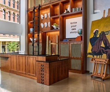 A modern luxury hotel lobby reception desk made of dark wood with a massive floor-to-ceiling wooden built-in bookshelf, a rolling library ladder, and a large oil painting of a trumpet player on an easel.