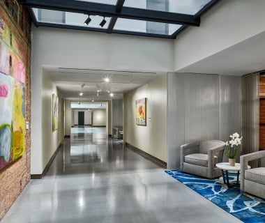 A long, modern hotel meeting hallway with polished concrete floors and large abstract paintings, featuring a seating area with two grey armchairs on a blue patterned rug under a glass skylight.