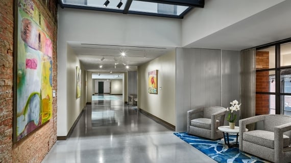 A long, modern hotel meeting hallway with polished concrete floors and large abstract paintings, featuring a seating area with two grey armchairs on a blue patterned rug under a glass skylight.