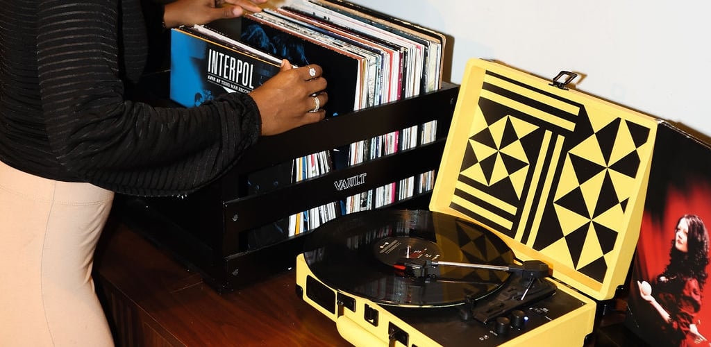 A person with dreadlocks browsing through a crate of vinyl records on a mid-century wooden console table next to a yellow portable record player.