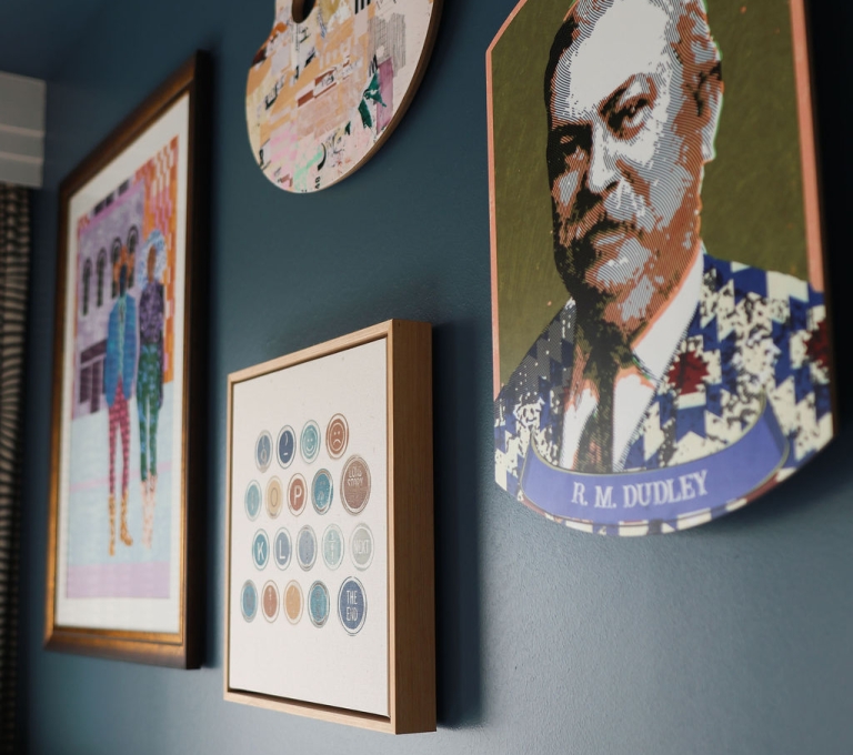 An eclectic gallery art wall featuring a stylized pixel-art style portrait of R. M. Dudley, a circular canvas, and framed prints mounted on a dark teal wall.
