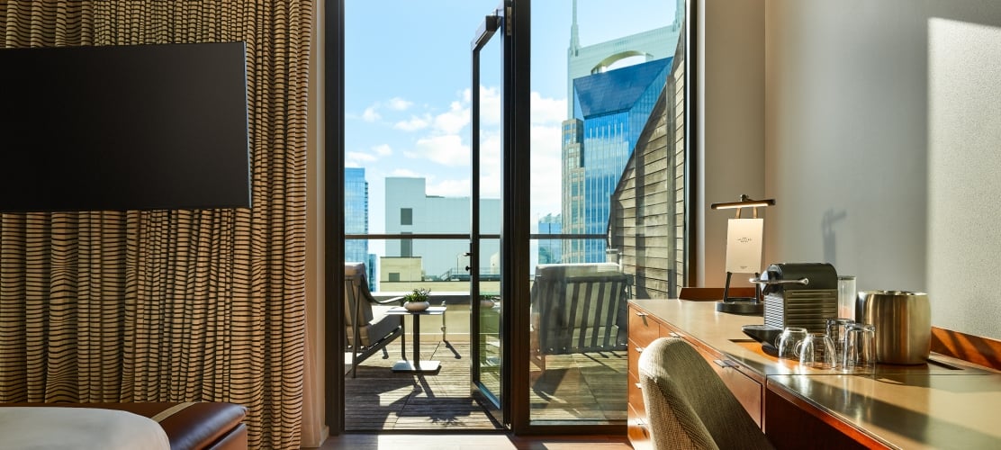 A modern hotel king room featuring a wooden desk and coffee station, looking out through open double glass doors onto a wooden terrace with outdoor seating and a view of the Nashville skyline.