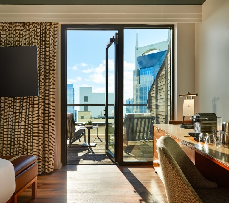 A modern hotel king room featuring a wooden desk and coffee station, looking out through open double glass doors onto a wooden terrace with outdoor seating and a view of the Nashville skyline.