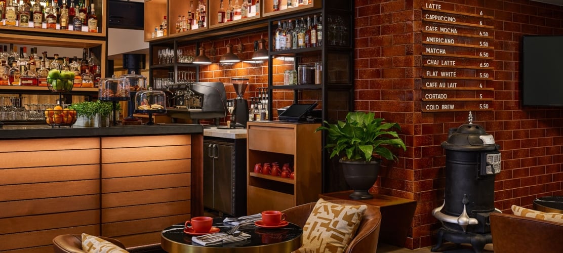 Cozy hotel lounge seating area with two leather armchairs and a small round table next to an exposed brick wall, a cafe menu board, and a lit bar counter in the background.