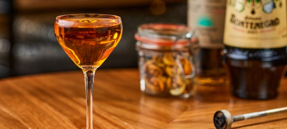 An amber cocktail served in a coupe glass on a wooden bar counter next to a lemon twist, a branded ice stamp, a jar of garnishes, and an Amaro Montenegro bottle.