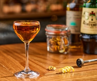 An amber cocktail served in a coupe glass on a wooden bar counter next to a lemon twist, a branded ice stamp, a jar of garnishes, and an Amaro Montenegro bottle.