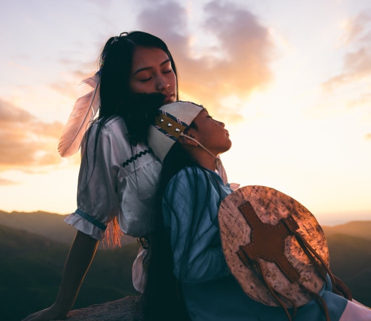 A child and mother in traditional Indigenous clothing, sitting closely together in a field under a vast sky
