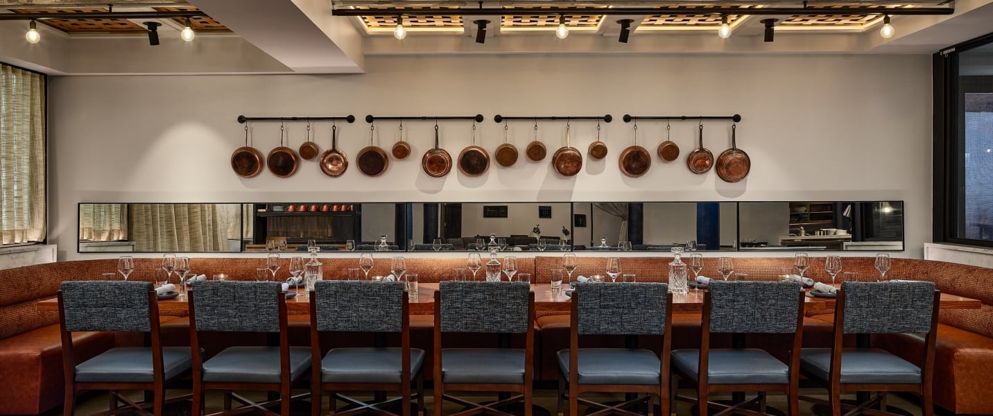 A long dining table is set for guests, flanked by a U-shaped booth. Copper pots hang above horizontal mirrors on the wall of the restaurant.
