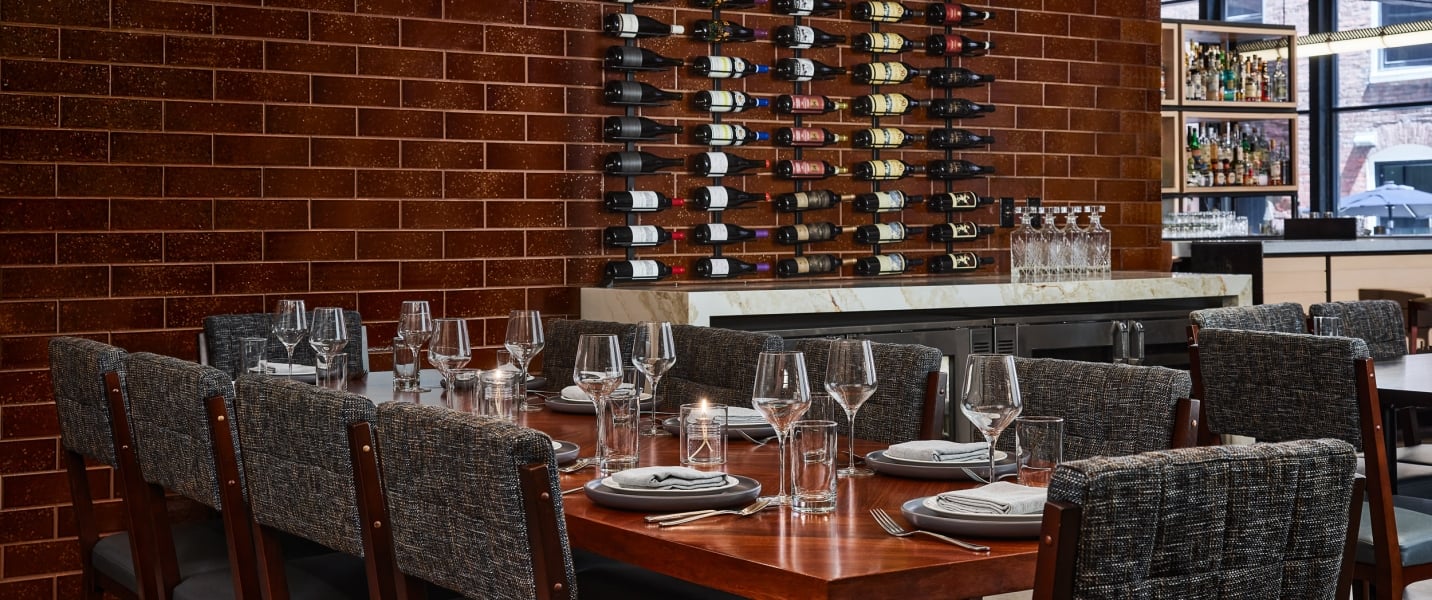 A long dining table is set with plates, glasses, and candles, facing a brown tiled wall featuring a large wine rack and a white marble bar counter.