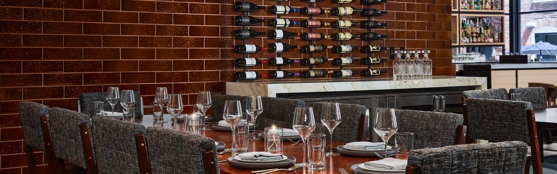 A long dining table is set with plates, glasses, and candles, facing a brown tiled wall featuring a large wine rack and a white marble bar counter.