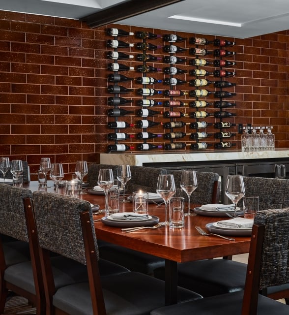 A long dining table is set with plates, glasses, and candles, facing a brown tiled wall featuring a large wine rack and a white marble bar counter.