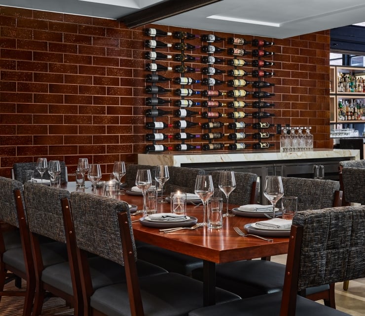 A long dining table is set with plates, glasses, and candles, facing a brown tiled wall featuring a large wine rack and a white marble bar counter.