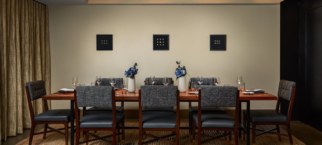 A long wooden dining table, set with chairs, dishes, and blue flowers, in a modern room featuring three square, nine-dot artworks on the wall.