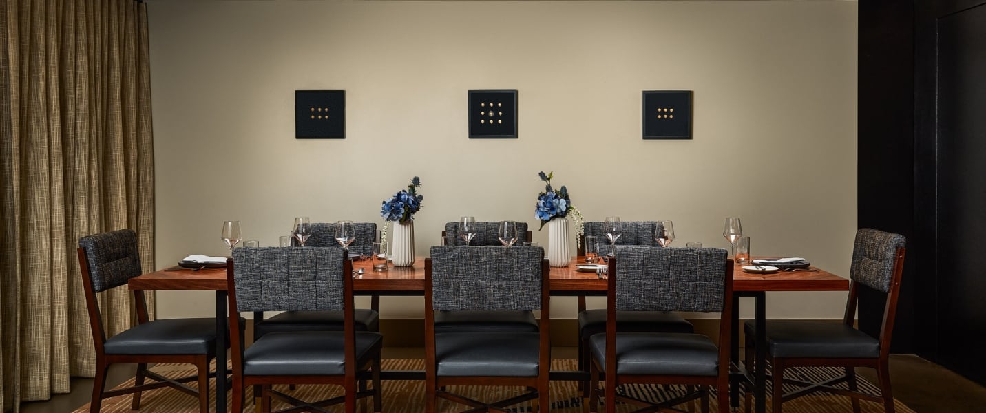 A long wooden dining table, set with chairs, dishes, and blue flowers, in a modern room featuring three square, nine-dot artworks on the wall.