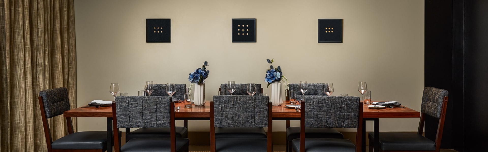 A long wooden dining table, set with chairs, dishes, and blue flowers, in a modern room featuring three square, nine-dot artworks on the wall.