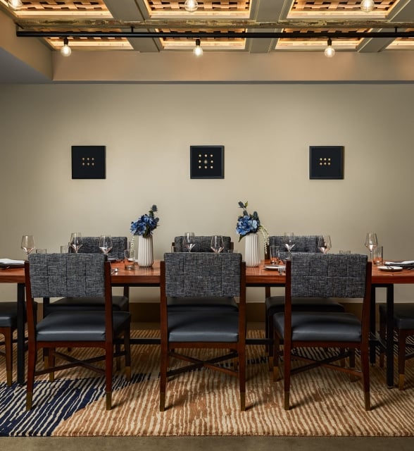 A long wooden dining table, set with chairs, dishes, and blue flowers, in a modern room featuring three square, nine-dot artworks on the wall.