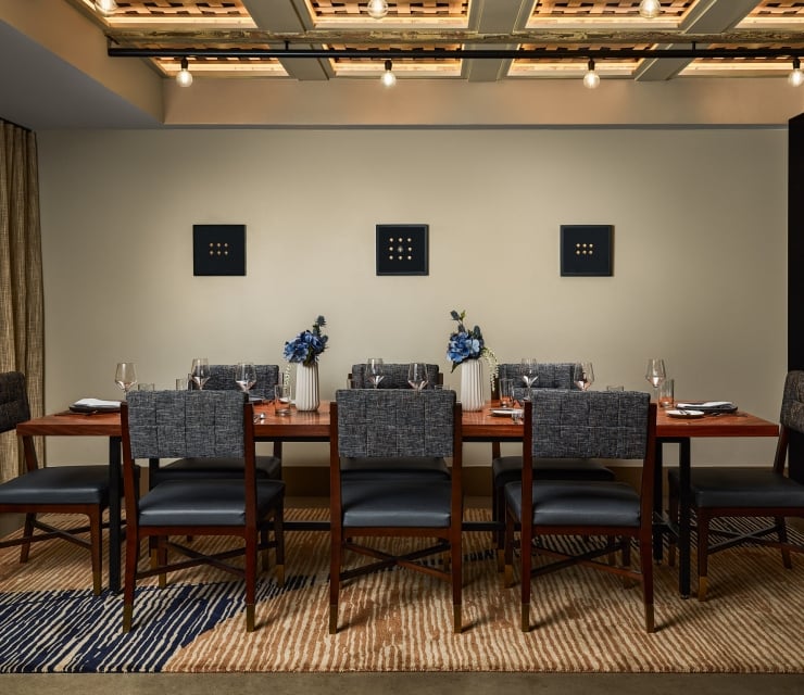 A long wooden dining table, set with chairs, dishes, and blue flowers, in a modern room featuring three square, nine-dot artworks on the wall.