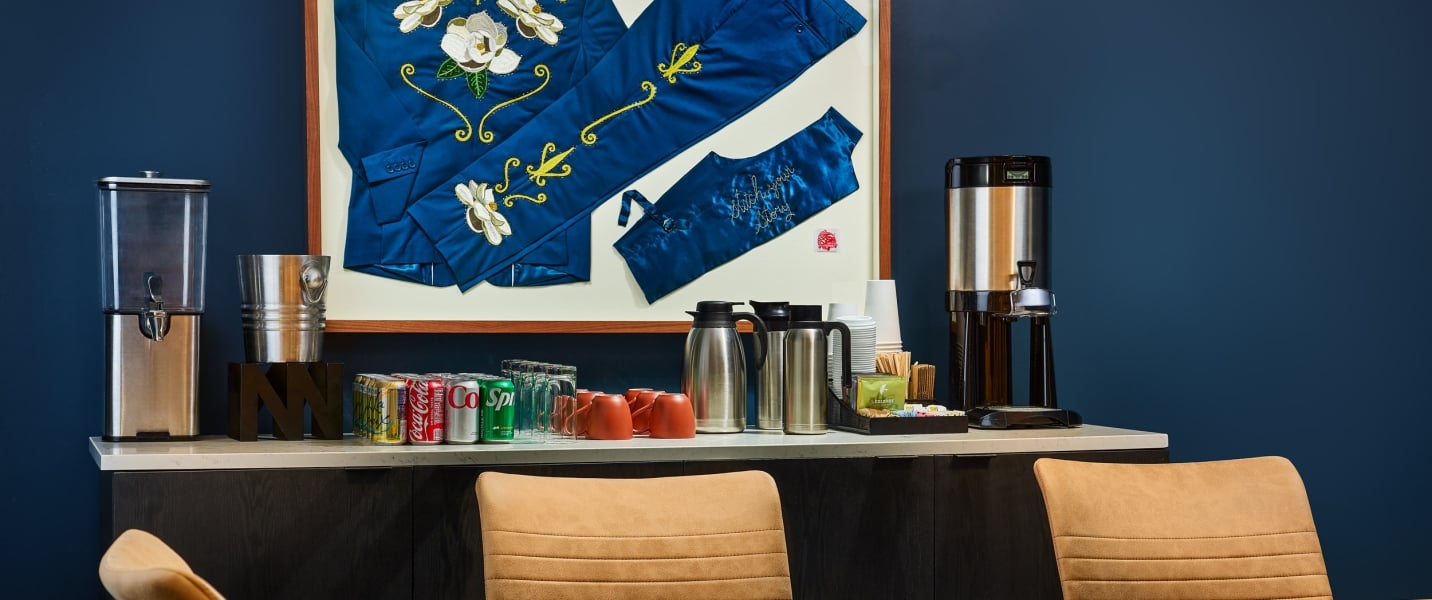 Beverage station offering sodas, coffee, and cups, is positioned under framed blue garments on a dark blue wall. One garment reads 'Stitch your story'.