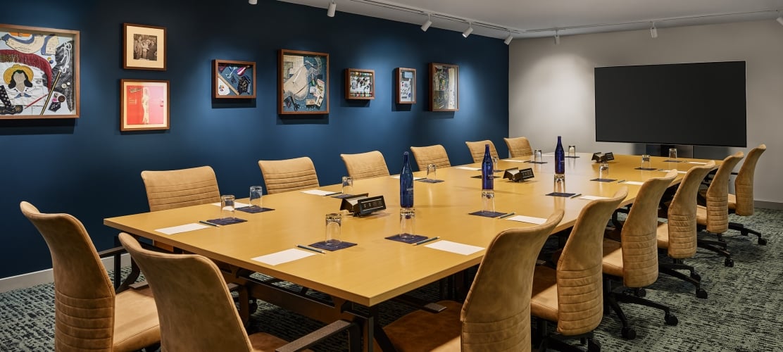 Long conference table with brown chairs set with Saint Géron water bottles, glasses, and papers. It's in a room with framed art on dark blue walls and a large TV.