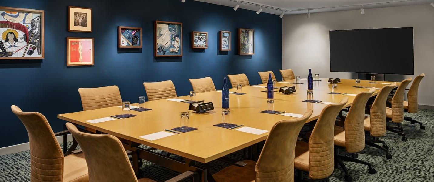 Long conference table with brown chairs set with Saint Géron water bottles, glasses, and papers. It's in a room with framed art on dark blue walls and a large TV.