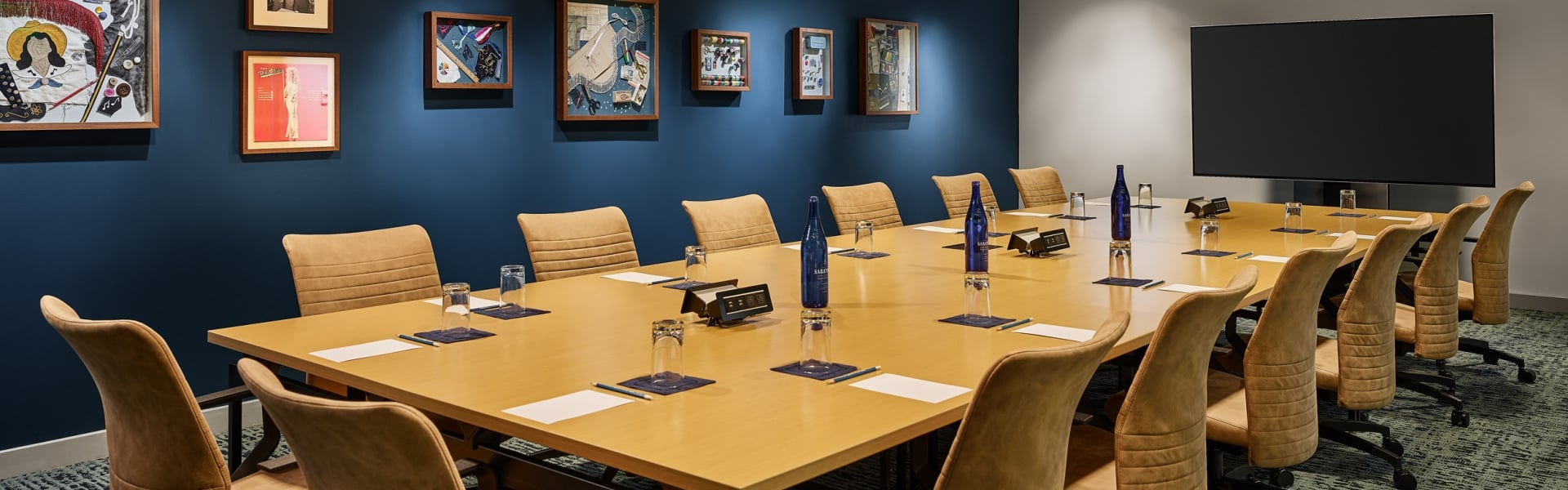 Long conference table with brown chairs set with Saint Géron water bottles, glasses, and papers. It's in a room with framed art on dark blue walls and a large TV.