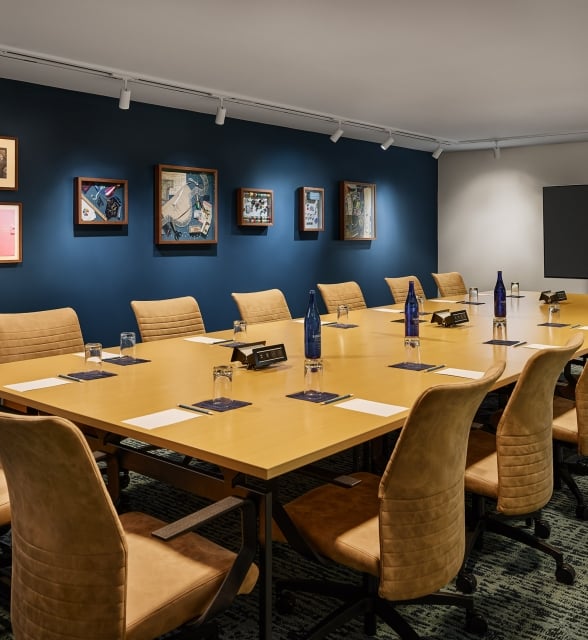 Long conference table with brown chairs set with Saint Géron water bottles, glasses, and papers. It's in a room with framed art on dark blue walls and a large TV.