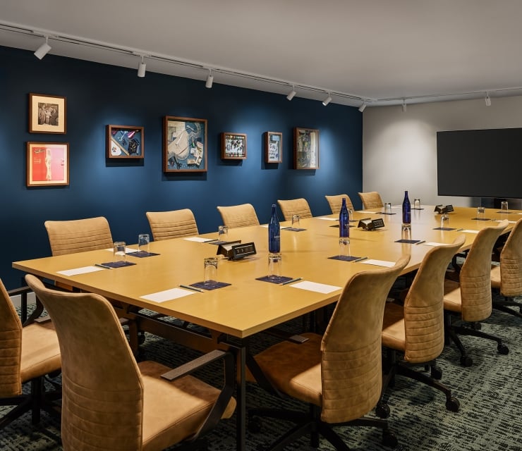 Long conference table with brown chairs set with Saint Géron water bottles, glasses, and papers. It's in a room with framed art on dark blue walls and a large TV.
