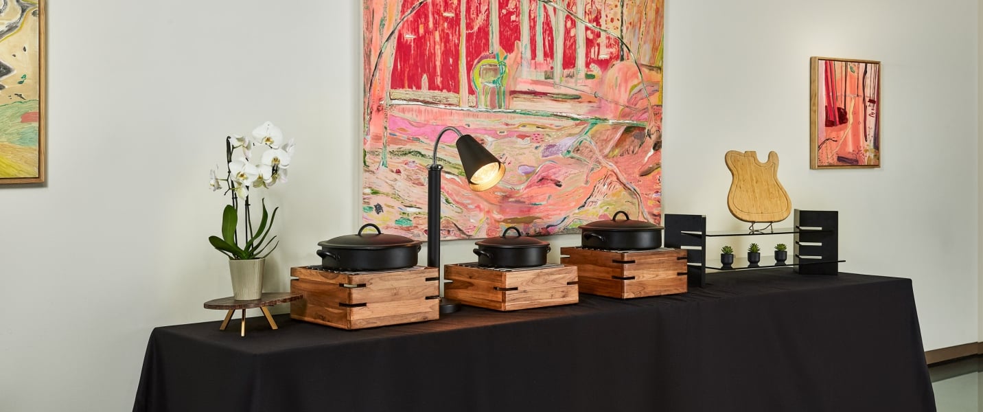 Three black chafing dishes rest on wooden warming stands, illuminated by a lamp, on a black-draped table with abstract art and an orchid nearby.