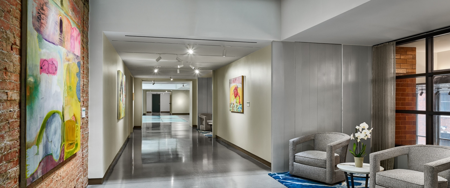 A modern hallway extends into the distance, featuring colorful abstract paintings on brick and plain walls, polished concrete floors, and a bright waiting area with armchairs.