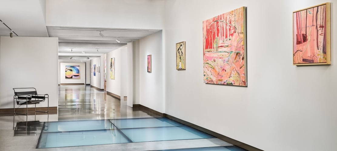 An art gallery hallway displays various colorful abstract paintings on white walls, featuring a light blue glass floor and a dark beamed ceiling.