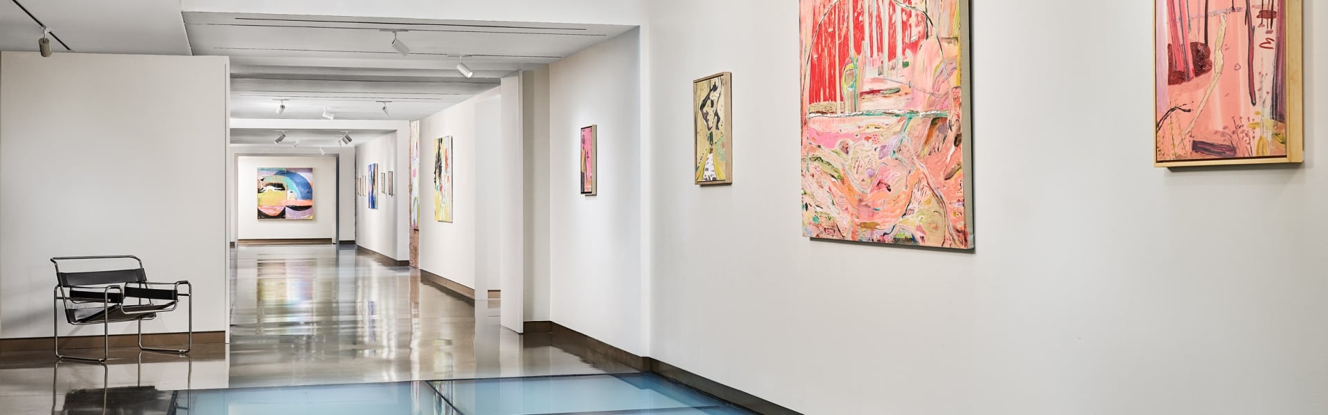 An art gallery hallway displays various colorful abstract paintings on white walls, featuring a light blue glass floor and a dark beamed ceiling.