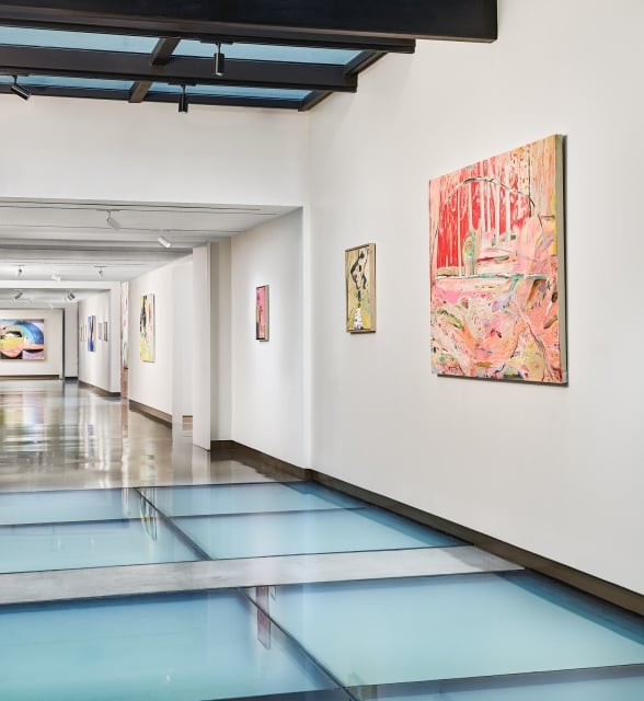 An art gallery hallway displays various colorful abstract paintings on white walls, featuring a light blue glass floor and a dark beamed ceiling.
