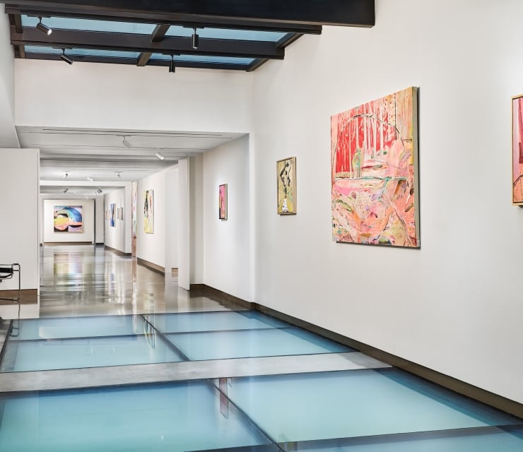 An art gallery hallway displays various colorful abstract paintings on white walls, featuring a light blue glass floor and a dark beamed ceiling.
