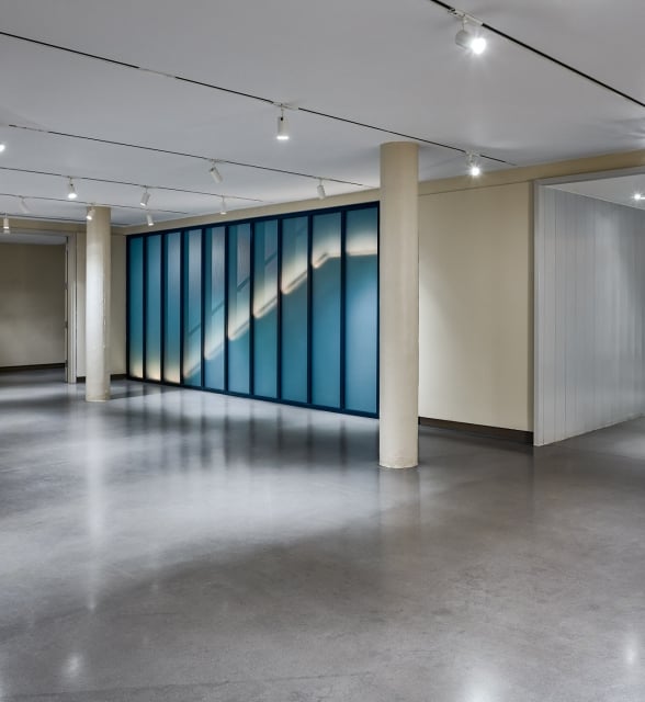 A long, blue-framed glass wall displays an illuminated diagonal pattern within a brightly lit modern interior space with a polished concrete floor.