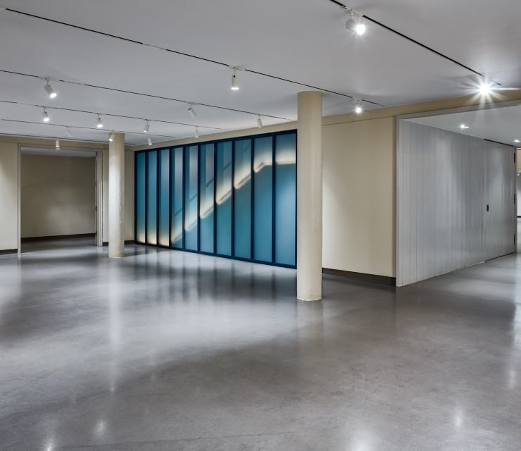 A long, blue-framed glass wall displays an illuminated diagonal pattern within a brightly lit modern interior space with a polished concrete floor.