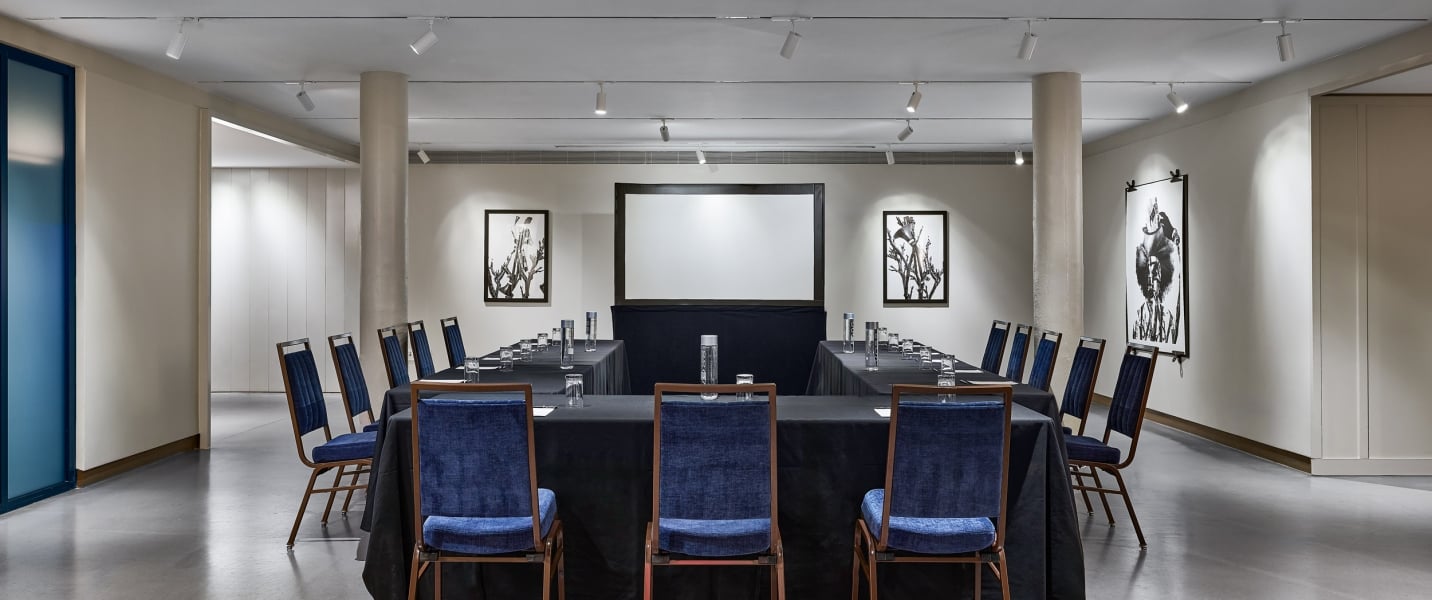 A U-shaped conference table is set with dark chairs, water, and glasses for a meeting within a modern room featuring a projection screen, abstract art, and track lighting.