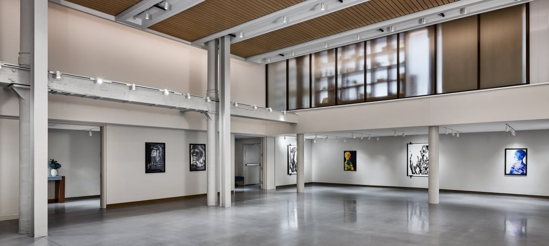 An empty modern art gallery displays various framed artworks on white walls, featuring a polished concrete floor, track lighting, and a slatted wood ceiling.